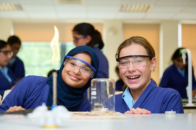 Two secondary-school girls wearing safety goggles smile as they carry out a science experiment in a laboratory classroom, with a Bunsen burner lit on the bench between them